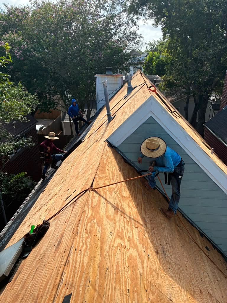 Three crew members working a steep plywood-decked roof around a dormer